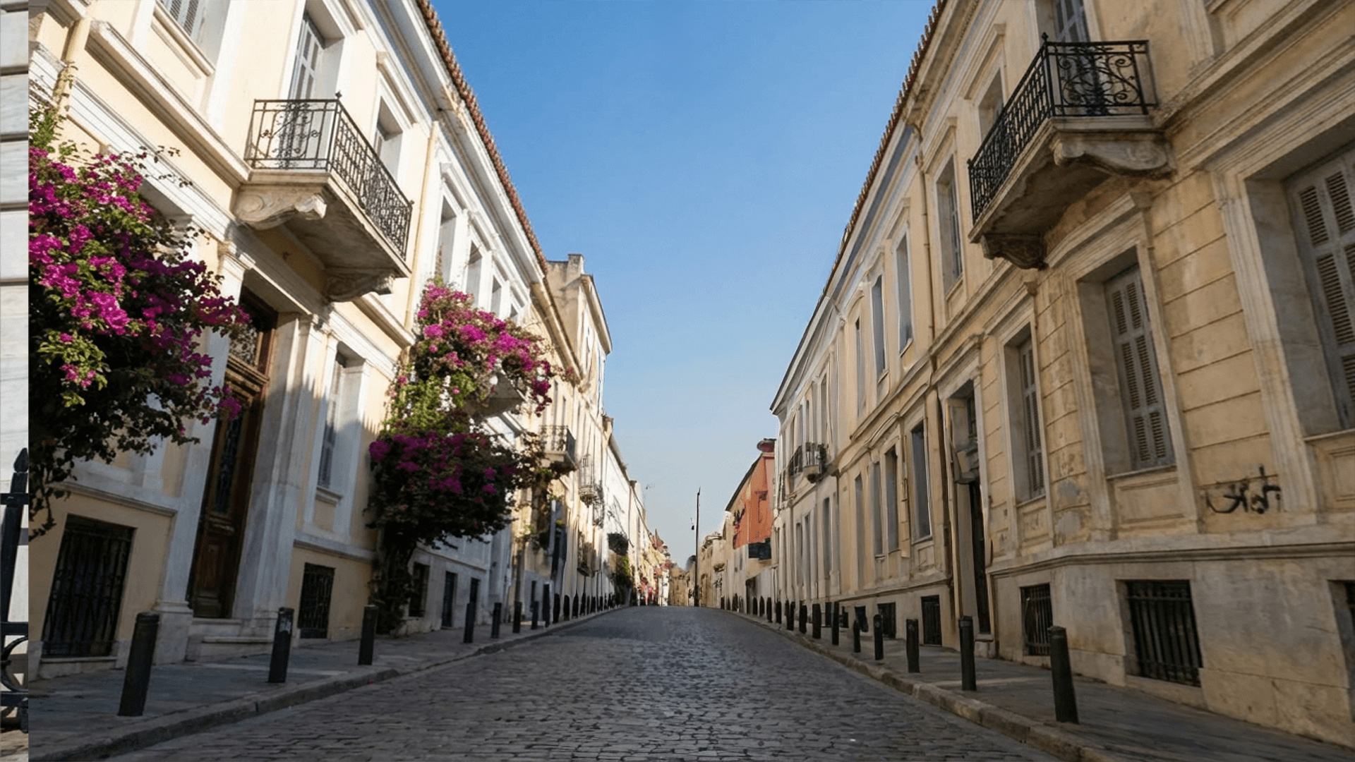 A quiet, narrow cobblestone street in Athens is lined with tall, historic buildings featuring balconies and blooming bougainvillea flowers, under a clear blue sky.